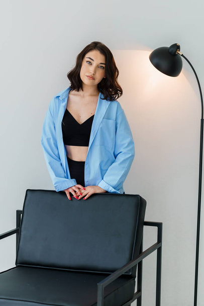 Beautiful girl in black trousers and a blue jacket near a chair on a white background. Portrait of a beautiful young woman - Foto, Bild