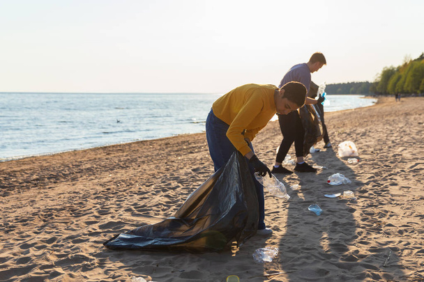 День Земли. Активисты-добровольцы собирают мусор для очистки береговой зоны. Женщины и мужчины кладут пластиковый мусор в мешок для мусора на берегу океана. Очистка прибрежной зоны - Фото, изображение
