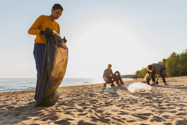 Dzień Ziemi. Wolontariusze zbierają śmieci w strefie przybrzeżnej plaży. Kobieta i mężczyzna wkładają plastikowe śmieci do worka na śmieci na brzegu oceanu. Ochrona środowiska czyszczenie stref przybrzeżnych - Zdjęcie, obraz
