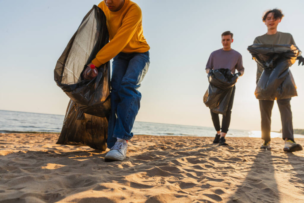 Dzień Ziemi. Zespół aktywistów wolontariuszy zbiera śmieci w strefie przybrzeżnej plaży. Kobieta wkłada plastikowe śmieci do worka na śmieci na brzegu oceanu. Ochrona środowiska czyszczenie stref przybrzeżnych - Zdjęcie, obraz
