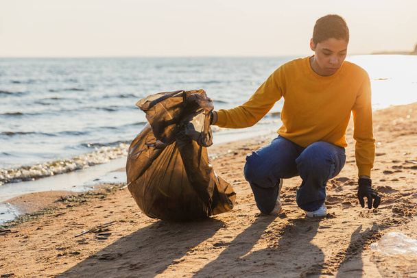 Jour de la Terre. L'équipe de militants bénévoles collecte les ordures ménagères de la zone côtière de la plage. Femme met des déchets en plastique dans un sac poubelle sur le rivage de l'océan. Conservation de l'environnement nettoyage des zones côtières - Photo, image
