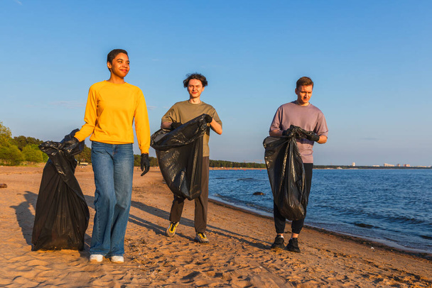 Dzień Ziemi. Zespół aktywistów wolontariuszy zbiera śmieci w strefie przybrzeżnej plaży. Kobieta ze śmieciami w worku na śmieci na brzegu oceanu. Ochrona środowiska czyszczenie stref przybrzeżnych - Zdjęcie, obraz