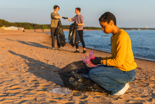 Día de la Tierra. Equipo de activistas voluntarios recoge la limpieza de la basura de la zona costera de la playa. La mujer pone basura plástica en la bolsa de basura en la orilla del océano. Limpieza de zonas costeras de conservación ambiental - Foto, Imagen