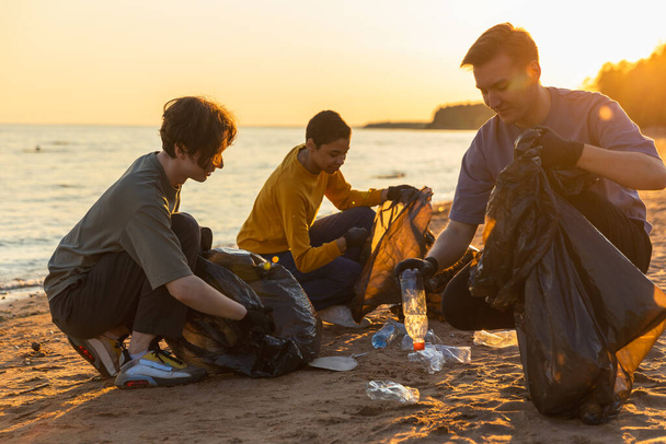Földi nap. Az önkéntesek aktivistái összegyűjtik a tengerparti övezet szemetének tisztítását. A férfiak műanyag szemetet tesznek a szemeteszsákba az óceán partján. Környezetvédelmi part menti övezetek tisztítása - Fotó, kép