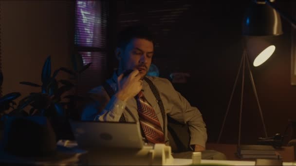 Μεσαία φορητή φωτογραφία του μπερδεμένου Καυκάσιου γενειοφόρου άνδρα σε ρετρό στυλ φορούν σκέψης κάθεται στο τραπέζι με σωλήνα στο χέρι τη νύχτα στο σκοτεινό δωμάτιο - Πλάνα, βίντεο