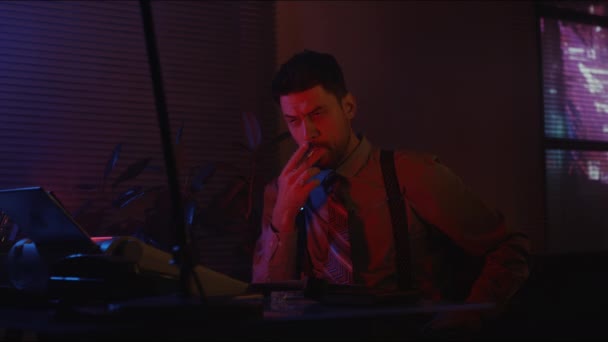 Medium side shot of Caucasian bearded man in old-fashioned vintage clothes smoking cigarette in neon lit office at night - Footage, Video