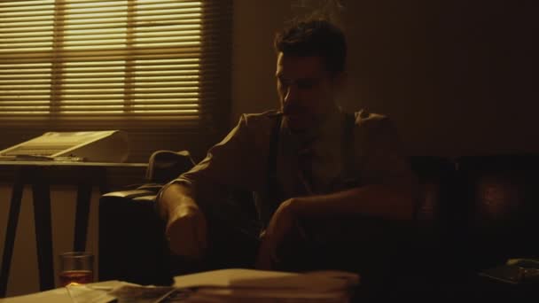 Střední sépiový snímek bělošského vousatého muže ve středním věku sedícího na pohovce, zapalujícího si cigaretu a házejícího fotku na stůl, který vypadal pesimisticky a otráveně, jak dává nohy na stůl v matně osvětleném pokoji - Záběry, video