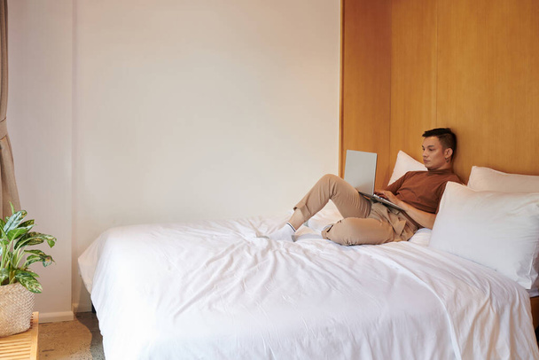 Vážné asijské mladý muž sedí na posteli a kódování na notebook - Fotografie, Obrázek