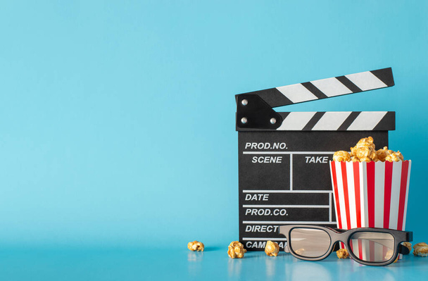 Experience excitement of much-awaited movie debut. Side view captures table scene with clapperboard, striped popcorn box, and 3D glasses against light blue wall, leaving space for text or film adverts - Photo, Image