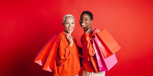 Portrait of a happy young female couple, wearing red sweater, carrying red shopping bags in front of a red background at studio. Two women together at studio. - Photo, Image
