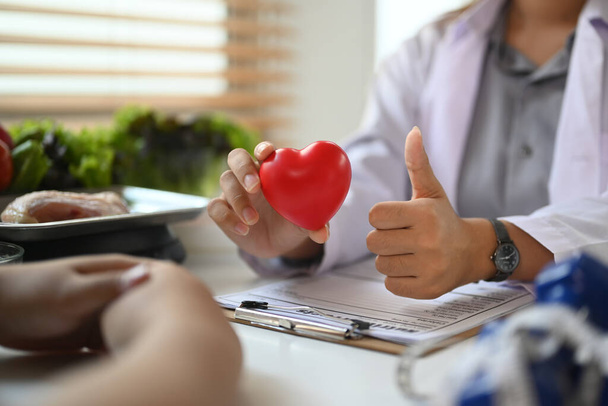 Klinikteki hastayla konuşurken beslenme uzmanı kırmızı kalbi formda tutuyor. Doğru beslenme ve sağlık bakımı kavramı. - Fotoğraf, Görsel