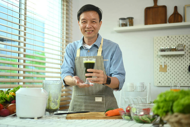 Retrato de homem sênior sorridente segurando vidro de smoothie verde em pé na cozinha. Conceito de estilo de vida saudável - Foto, Imagem