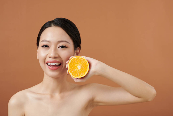 Ευτυχισμένη νεαρή γυναίκα με αψεγάδιαστη επιδερμίδα που κρατάει το μισό πορτοκάλι - Φωτογραφία, εικόνα