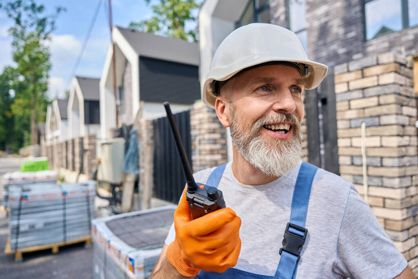 Cheerful builder with walkie-talkie in hand standing on street among building materials - Foto, afbeelding