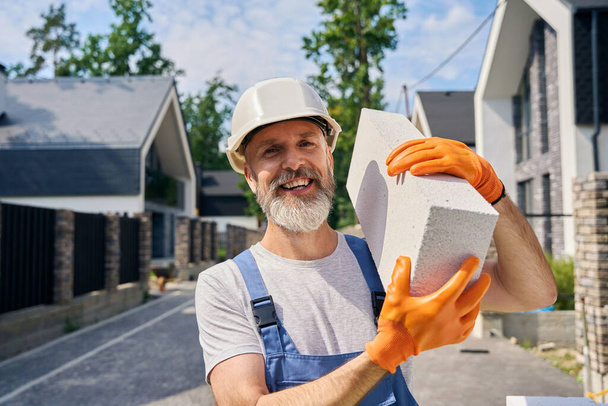 Smiling construction worker in rubber gloves holding concrete paver block with both hands - Foto, afbeelding