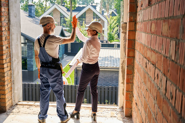 Cheerful building inspector giving high-five to contented builder while standing inside unfinished residential house - Foto, Imagem
