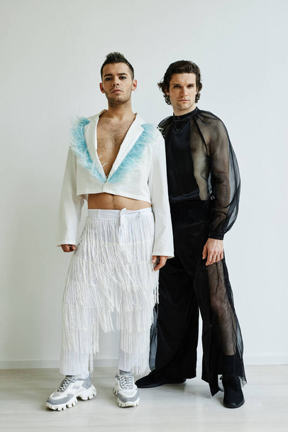 Vertical full length portrait of two non binary young men wearing extravagant outfits standing against white wall in fashion studio - Photo, Image