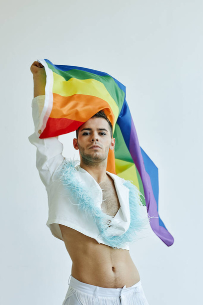 Vertical portrait of young gay man waving pride flag and wearing crop top against white - Photo, Image