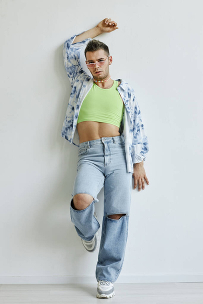 Vertical full length portrait of extravagant young gay man leaning against white wall in fashion studio - Photo, Image