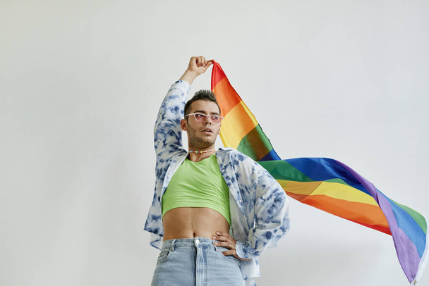 Retrato de bajo ángulo del hombre gay confiado ondeando bandera de orgullo contra fondo blanco, espacio de copia - Foto, imagen