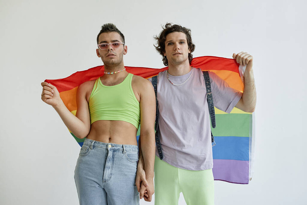 Portrait of two gay young men holding hands with pride flag against white background - Фото, изображение