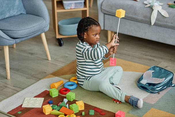 Портрет в полный рост симпатичного черного ребенка, играющего с игрушками на полу в уютной домашней обстановке, пространство для копирования - Фото, изображение