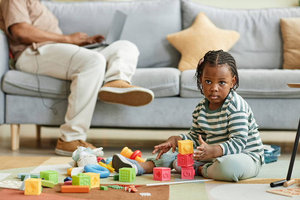 Retrato de longitud completa de lindo niño negro jugando con bloques de juguete mientras está sentado en el suelo en casa con el padre trabajando en segundo plano, espacio de copia - Foto, Imagen