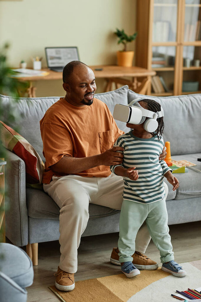 Вертикальный портрет милого черного ребенка в наушниках VR и наслаждающегося развлечениями виртуальной реальности при поддержке отца - Фото, изображение