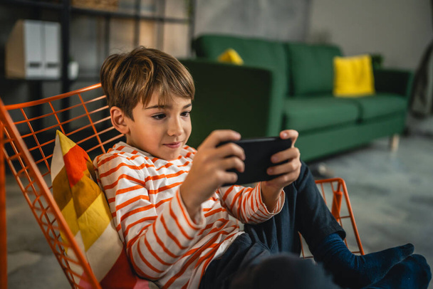 Bir çocuk evde oturuyor cep telefonunu tutuyor akıllı telefon video oyunları oynuyor - Fotoğraf, Görsel