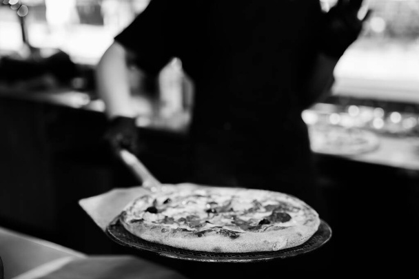 Imagen en blanco y negro mostrando una pizza artesanal recién horneada con un chef borroso en el fondo. - Foto, imagen