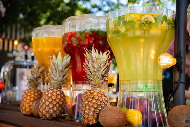 Een reeks kleurrijke, met fruit doordrenkte waterdispensers, geflankeerd door ananas en kokosnoten, creëren een tropische verfrissingscène - Foto, afbeelding