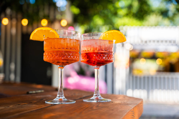 Twee sprankelende aperitief cocktails met oranje garnering, op een houten bar oppervlak, met een bokeh verlichting achtergrond - Foto, afbeelding