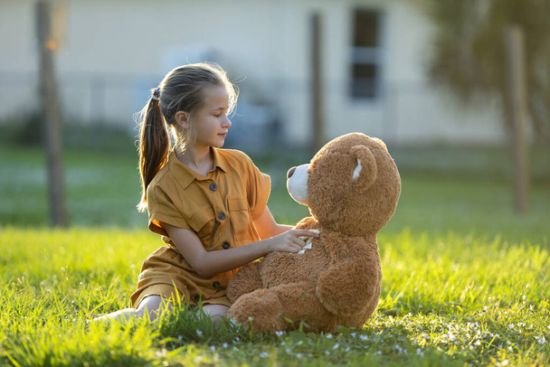 Güzel bir kız çocuğu oyuncak ayısı arkadaşını yeşil çimlerin üzerinde nazikçe kucaklıyor. Arkadaşlık değerleri kavramı. - Fotoğraf, Görsel