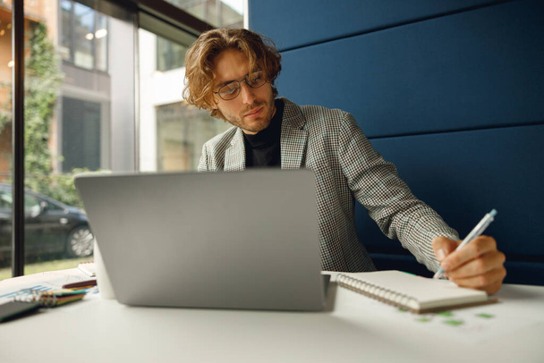 Красивый бизнесмен, работающий над проектом, используя ноутбук и делая заметки, сидя в офисе - Фото, изображение