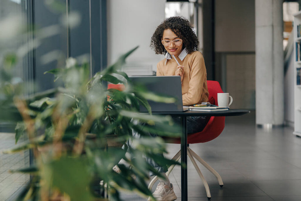 Stylová obchodní žena v brýlích pracuje na notebooku, zatímco sedí v moderní kanceláři v blízkosti okna - Fotografie, Obrázek