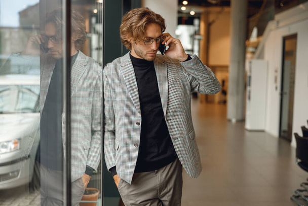 Handsome male freelancer in eyeglasses is talking phone standing in modern coworking space - Photo, Image