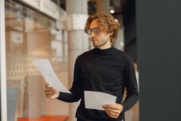 Όμορφος επιχειρηματίας κοιτάζοντας τα έγγραφα, ενώ στέκεται στο σύγχρονο φόντο του γραφείου - Φωτογραφία, εικόνα