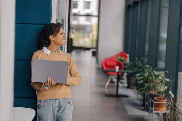 Jovem mulher de negócios sorrindo usando laptop enquanto está de pé no moderno escritório fundo - Foto, Imagem