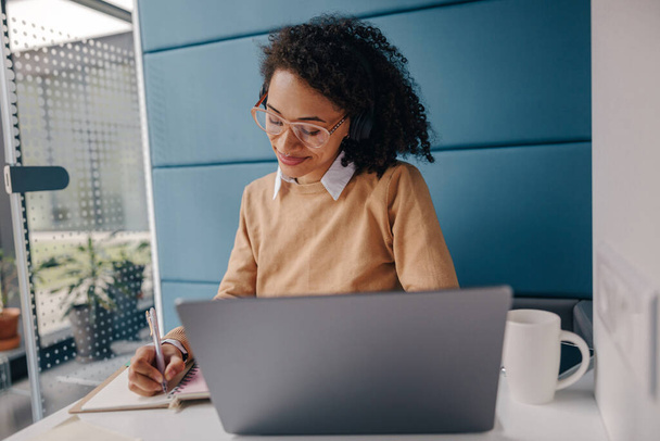 Femme entrepreneure souriante travaillant sur un ordinateur portable, prenant des notes dans un espace de coworking confortable intérieur - Photo, image