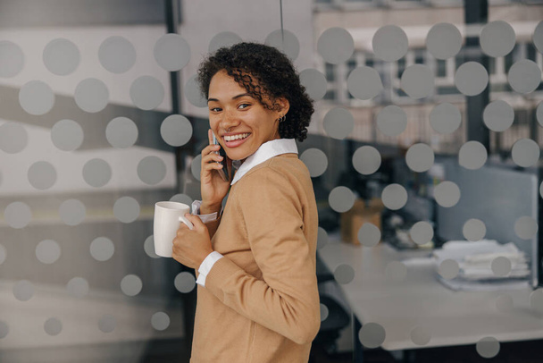 Χαμογελώντας γυναίκα διευθυντής με φλιτζάνι καφέ μιλάει τηλέφωνο, ενώ στέκεται στο σύγχρονο φόντο του γραφείου  - Φωτογραφία, εικόνα
