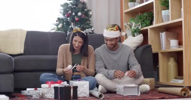 Kerstmis, koppel en cadeauverpakking in huis met papier en feest met feestmuts. Happy, conversatie en jongeren met cadeau, doos en communicatie in een woonkamer voor vakantie met liefde. - Video