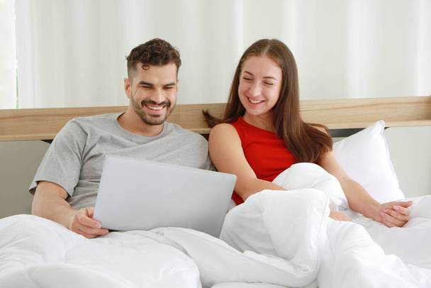 Lächelndes kaukasisches Paar, das sich entspannt und gemeinsam auf dem Laptop in den sozialen Medien im Schlafzimmer einen lustigen Film anschaut. Arbeit von zu Hause aus und Lebensstil im Urlaub - Foto, Bild