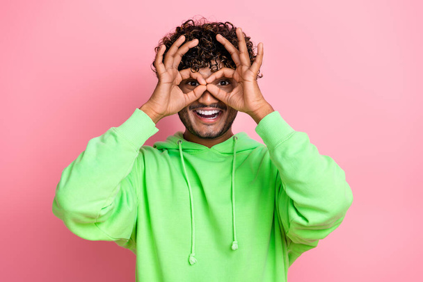 Portrait d'un jeune étudiant funky portant un sweat-shirt vert élégant montrant un symbole de jumelles sur des yeux isolés sur un fond rose. - Photo, image