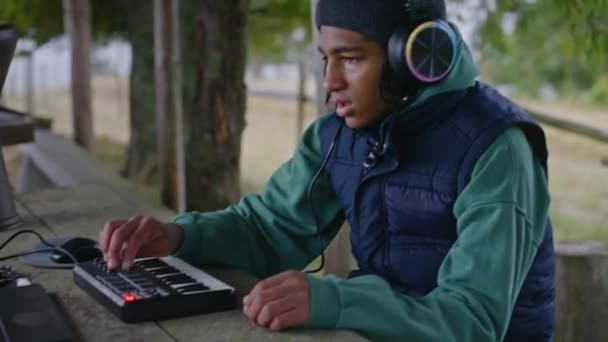 Adolescente afroamericano con auriculares juega en el controlador MIDI al aire libre. Joven músico, compositor crea música, mezcla canciones, utiliza portátil para grabar mientras está sentado en el cenador en el bosque. - Imágenes, Vídeo