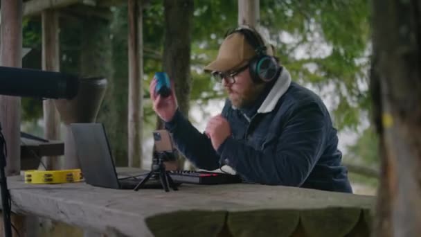ヘッドフォンの男性ミュージシャンはMIDIキーボードで演奏し,シェーカーを使用して森の外に座っている音楽を構成します. コーカサス人の男は,山への休暇中にトリポッドでラップトップと電話で曲を記録します. - 映像、動画