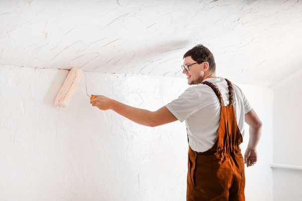 Kleur van het jaar 2024: Perzik Fuzz. Reparatie, woningverbetering en renovatie concept. jonge man in overall schilderen muur in kleur met een rol - Foto, afbeelding