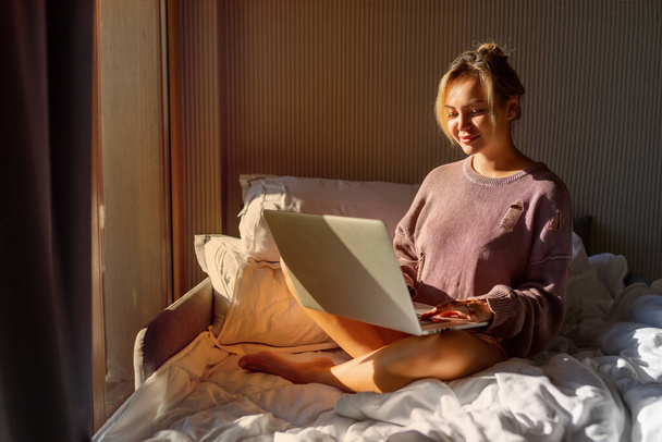 Ευτυχισμένη περιστασιακή όμορφη γυναίκα που εργάζεται σε ένα φορητό υπολογιστή κάθεται στο κρεβάτι στο σπίτι - Φωτογραφία, εικόνα