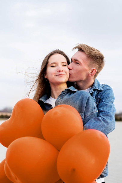 Color del año 2024: Melocotón Fuzz. Día de San Valentín. Joven pareja amorosa abrazando y sosteniendo globos en forma de corazón al aire libre - Foto, imagen