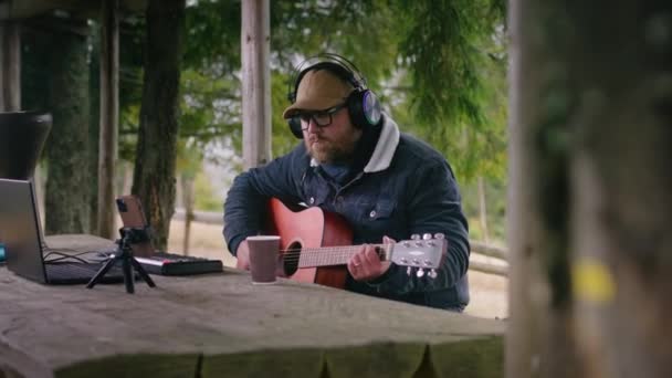 Kaukasischer Mann mit Kopfhörern spielt Gitarre in einem hölzernen Pavillon im Wald. Profimusiker erstellt und nimmt während einer Urlaubsreise in den Bergen Musik mit Laptop und Handy auf Stativ auf. - Filmmaterial, Video