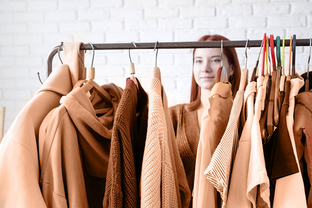 Цвет года 2024: Персиковый пушок. красивая молодая женщина в бежевом пальто, стоящая перед вешалкой и выбирающая наряды. Выбор гардероба, стилист, шоппинг. Проектирование одежды - Фото, изображение